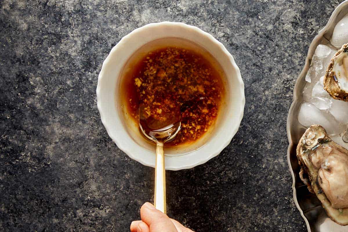 Bowl of soy sauce and ginger mixture being stirred with a spoon next to oysters on ice.