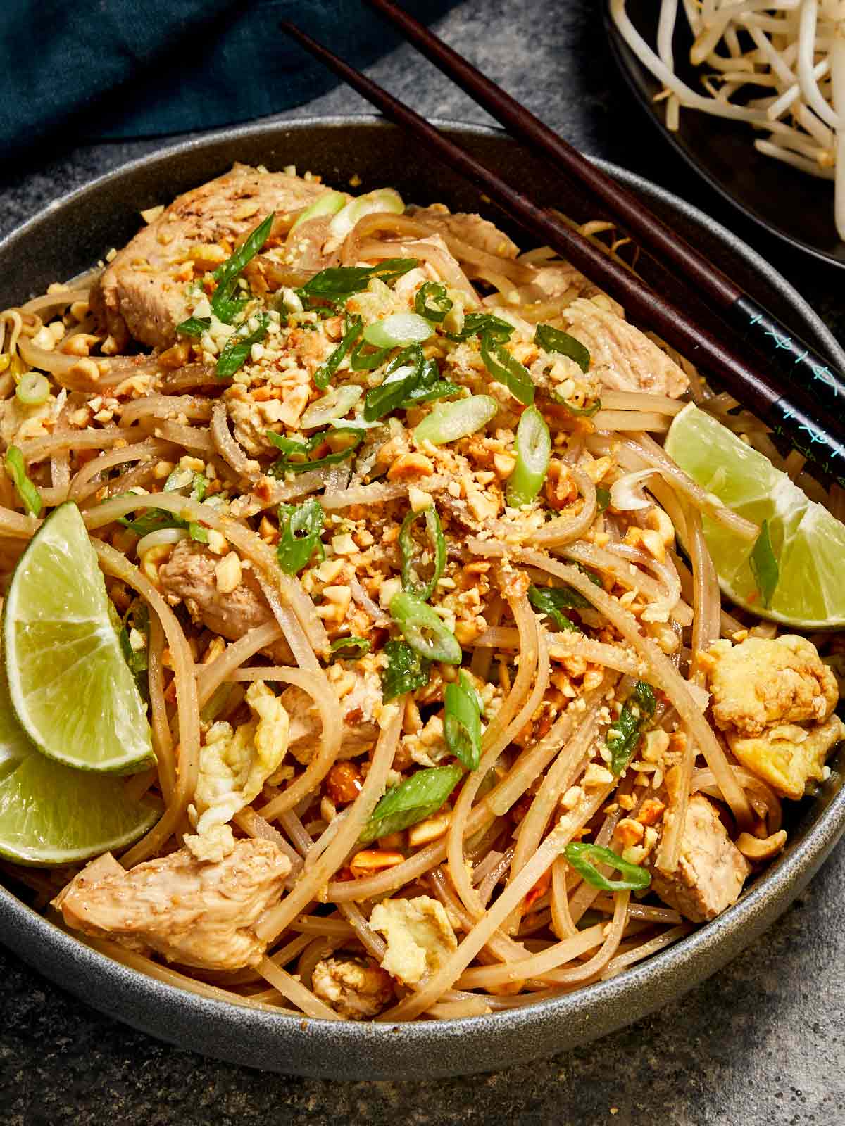 Bowl of Pad Thai with chicken, tofu, scrambled egg, bean sprouts, and peanuts, garnished with lime wedges and served with chopsticks.
