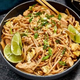 Bowl of Pad Thai with chicken, tofu, scrambled egg, bean sprouts, and crushed peanuts, garnished with lime wedges and green onions, served with chopsticks.