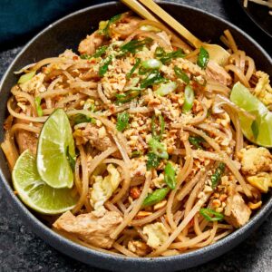 Bowl of Pad Thai with chicken, tofu, scrambled egg, bean sprouts, and crushed peanuts, garnished with lime wedges and green onions, served with chopsticks.