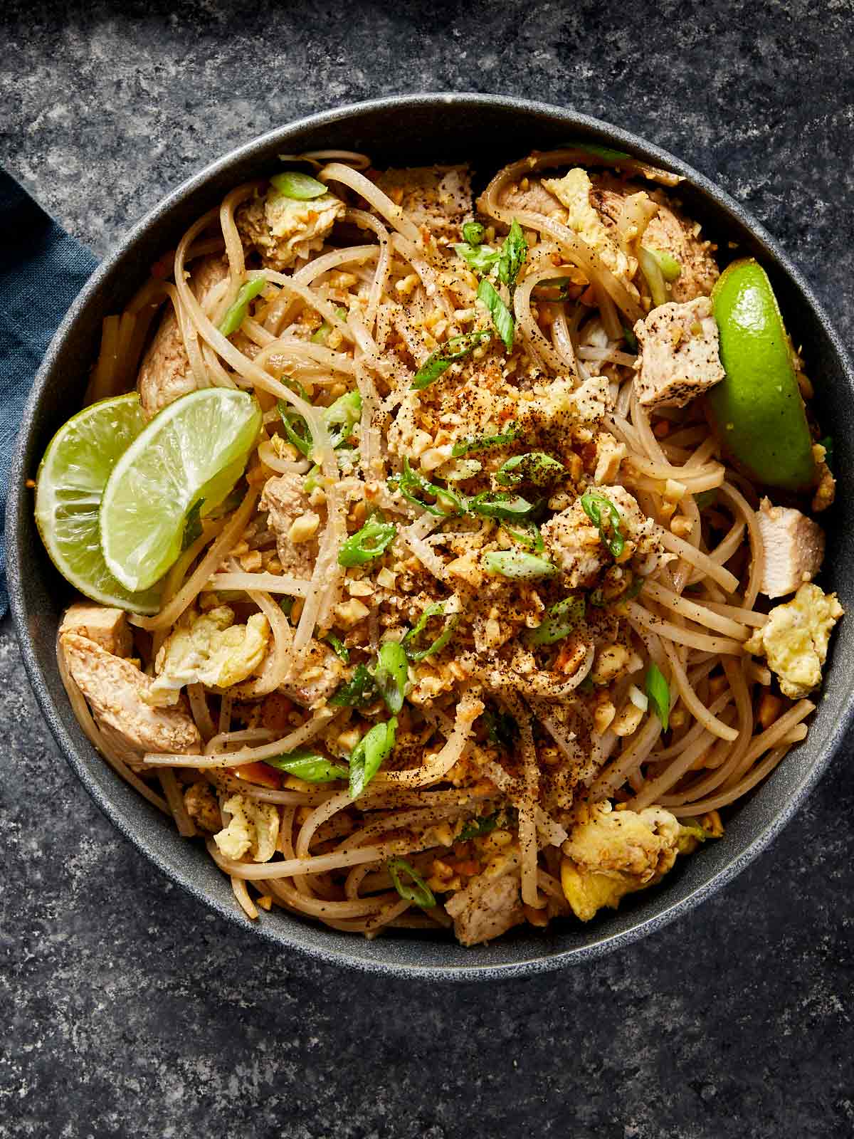 Overhead view of a bowl of chicken Pad Thai topped with scrambled egg, bean sprouts, crushed peanuts, green onions, and lime wedges.