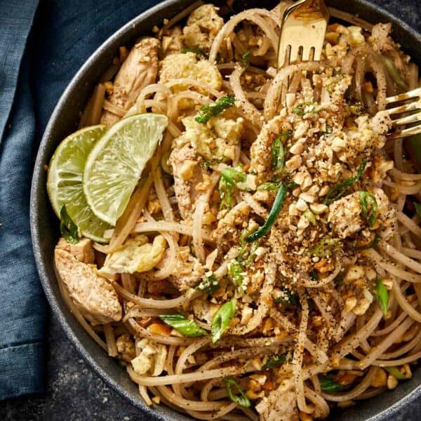 Bowl of chicken Pad Thai topped with crushed peanuts, green onions, and lime wedges, served with a golden fork on a dark background.