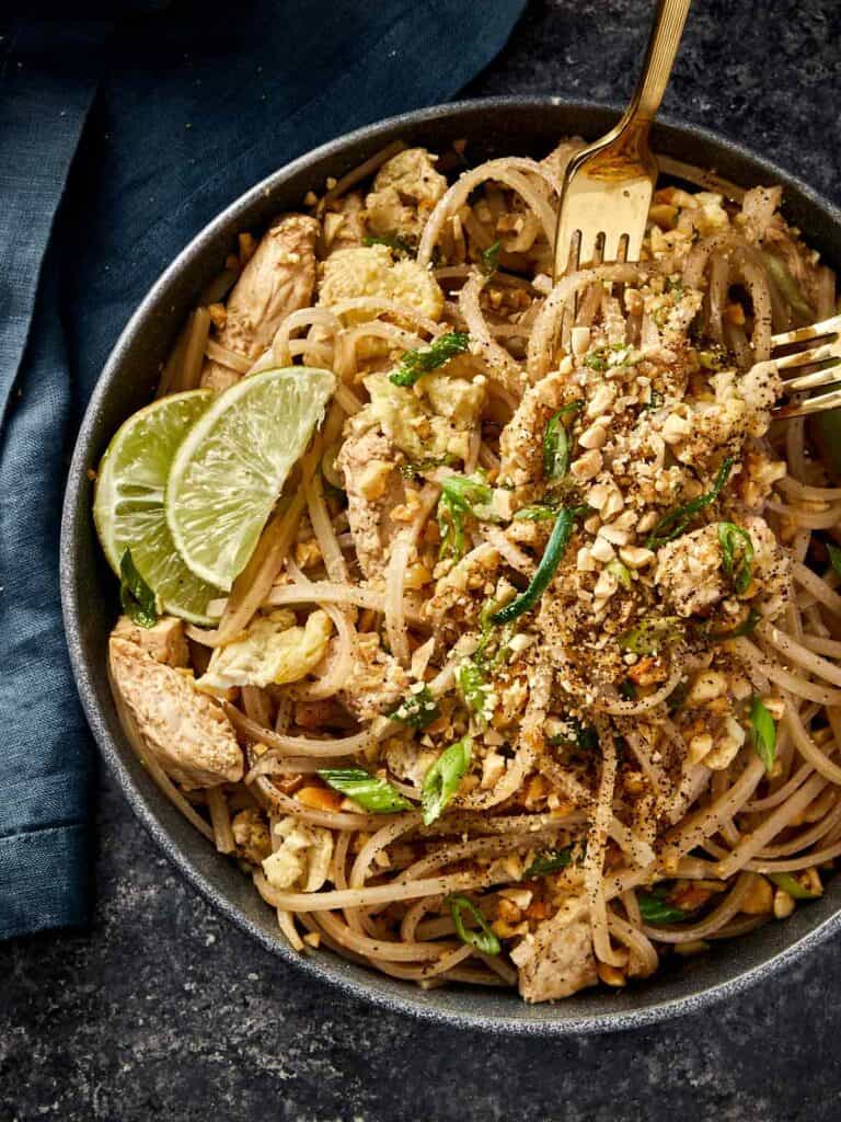 Bowl of chicken Pad Thai topped with crushed peanuts, green onions, and lime wedges, served with a golden fork on a dark background.