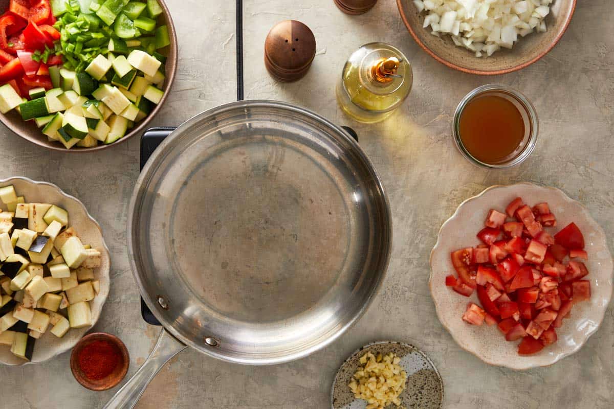 Empty skillet surrounded by bowls of diced eggplant, zucchini, tomatoes, peppers, onion and garlic, plus oil and broth.