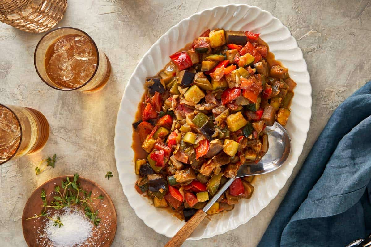 Ratatouille served on an oval platter with a spoon, surrounded by drinks and herbs.