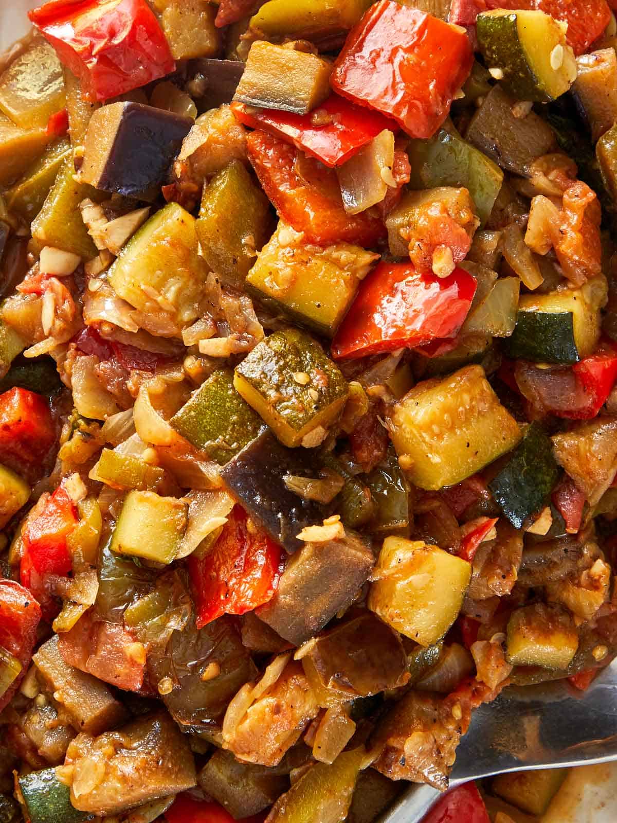 A vibrant bowl of ratatouille, filled with assorted cooked vegetables, accompanied by a spoon for serving.