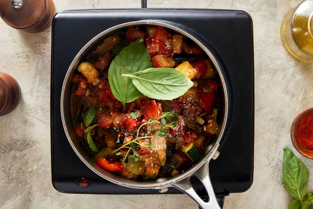 Pot of ratatouille simmering on a stovetop with fresh herbs on top.