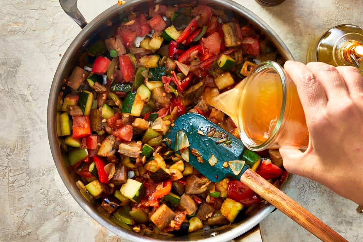 Broth being poured over sautéed vegetables in a skillet.