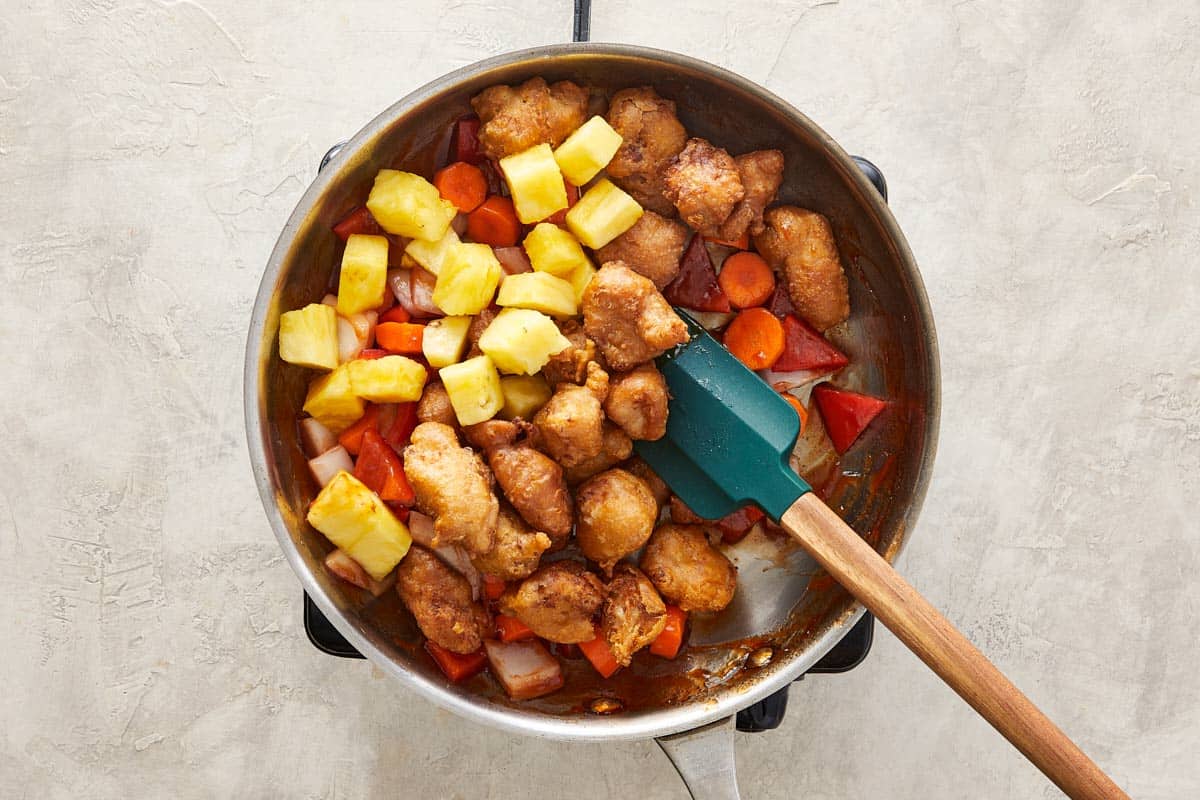 Wok with fried chicken, pineapple, carrots, onion, and bell pepper mixed with sauce.