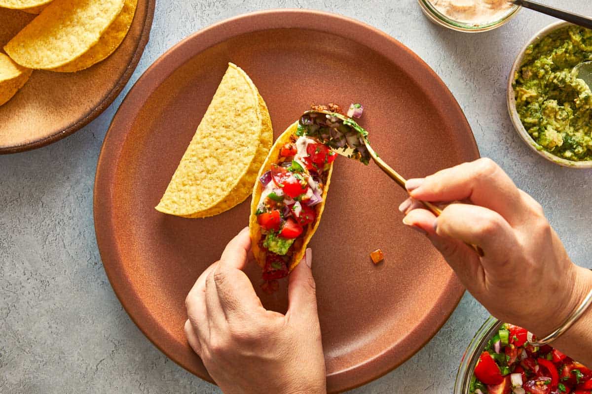 Hands filling a taco shell with beef, guacamole, and pico de gallo