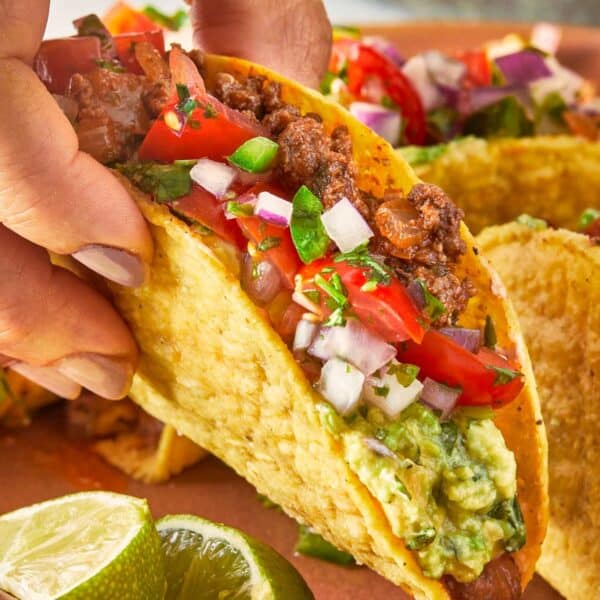 Hand holding a beef taco with guacamole, salsa, and lime on the side
