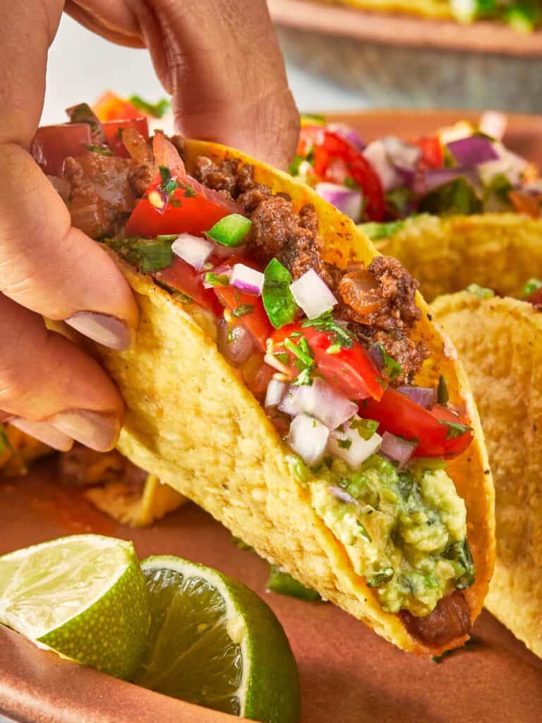 Hand holding a beef taco with guacamole, salsa, and lime on the side