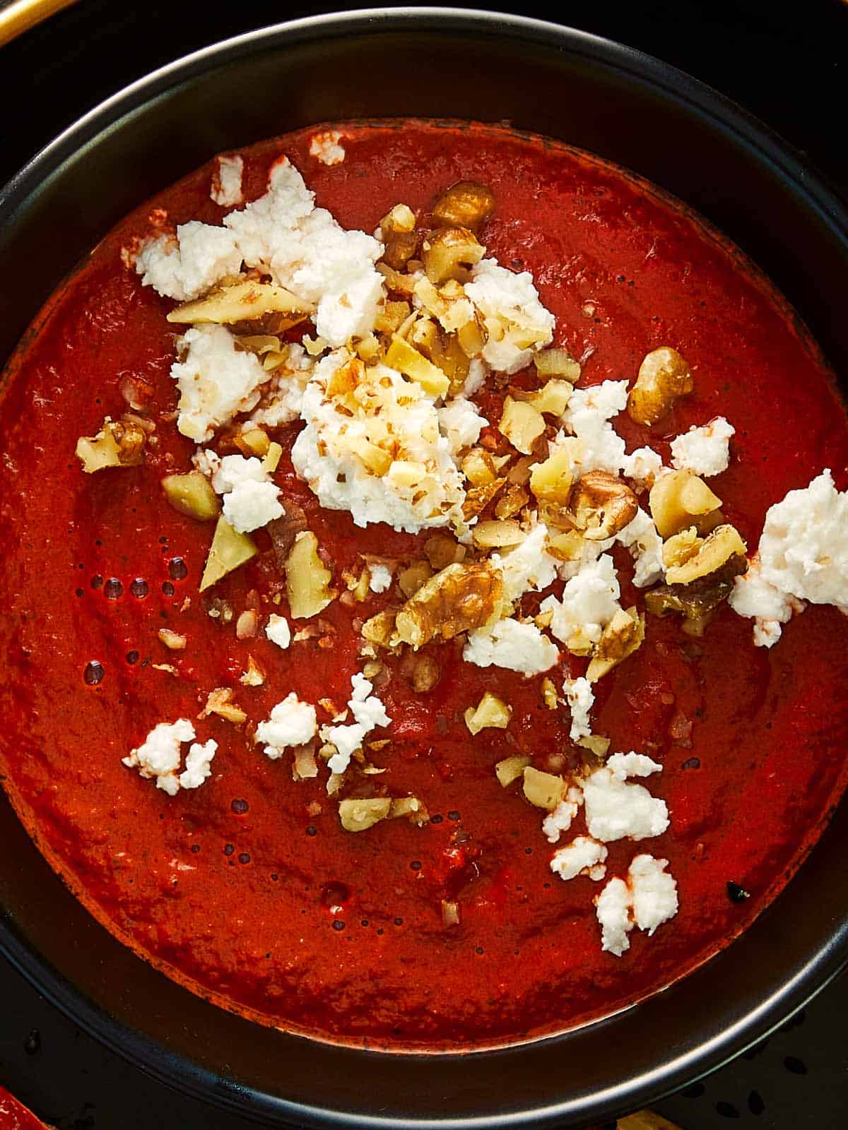 Close-up of a rich, vibrant red dip topped with crumbled feta cheese and chopped walnuts, served in a black bowl. The creamy, smoky texture and contrast of colors give this Bloody Dip an appetizing Halloween vibe.