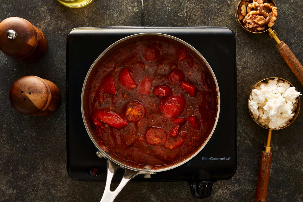 Simmering mixture of tomatoes and red peppers in a saucepan, thickened into a rich red sauce, with feta and walnuts on the side.