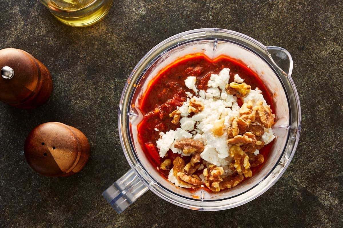 Blender filled with roasted red sauce, feta cheese, and walnuts ready to blend into a smooth, creamy dip.