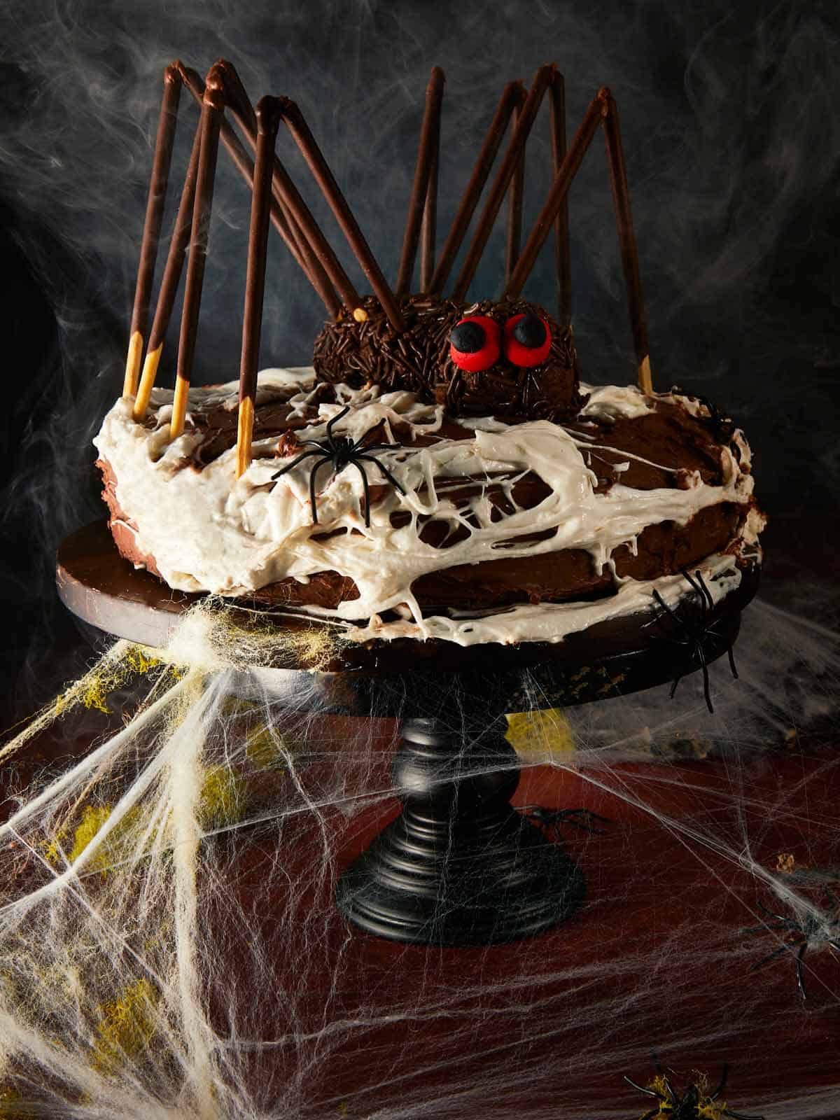 Halloween chocolate layer cake styled as a giant spider, surrounded by fake cobwebs and plastic spiders on a dark red table.