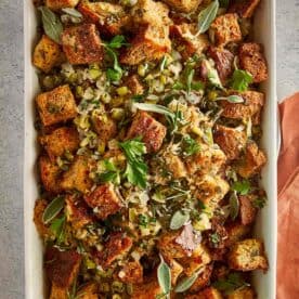 Overhead view of a rectangular baking dish filled with homemade herb stuffing, with toasted bread pieces, flecks of green herbs, and a gold serving spoon nestled inside.