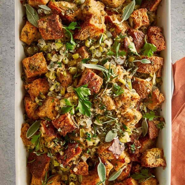 Overhead view of a rectangular baking dish filled with homemade herb stuffing, with toasted bread pieces, flecks of green herbs, and a gold serving spoon nestled inside.