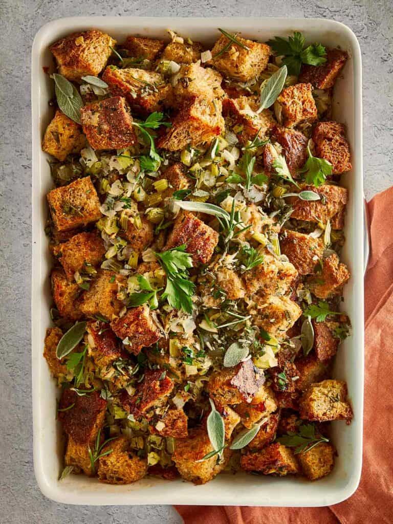 Overhead view of a rectangular baking dish filled with homemade herb stuffing, with toasted bread pieces, flecks of green herbs, and a gold serving spoon nestled inside.