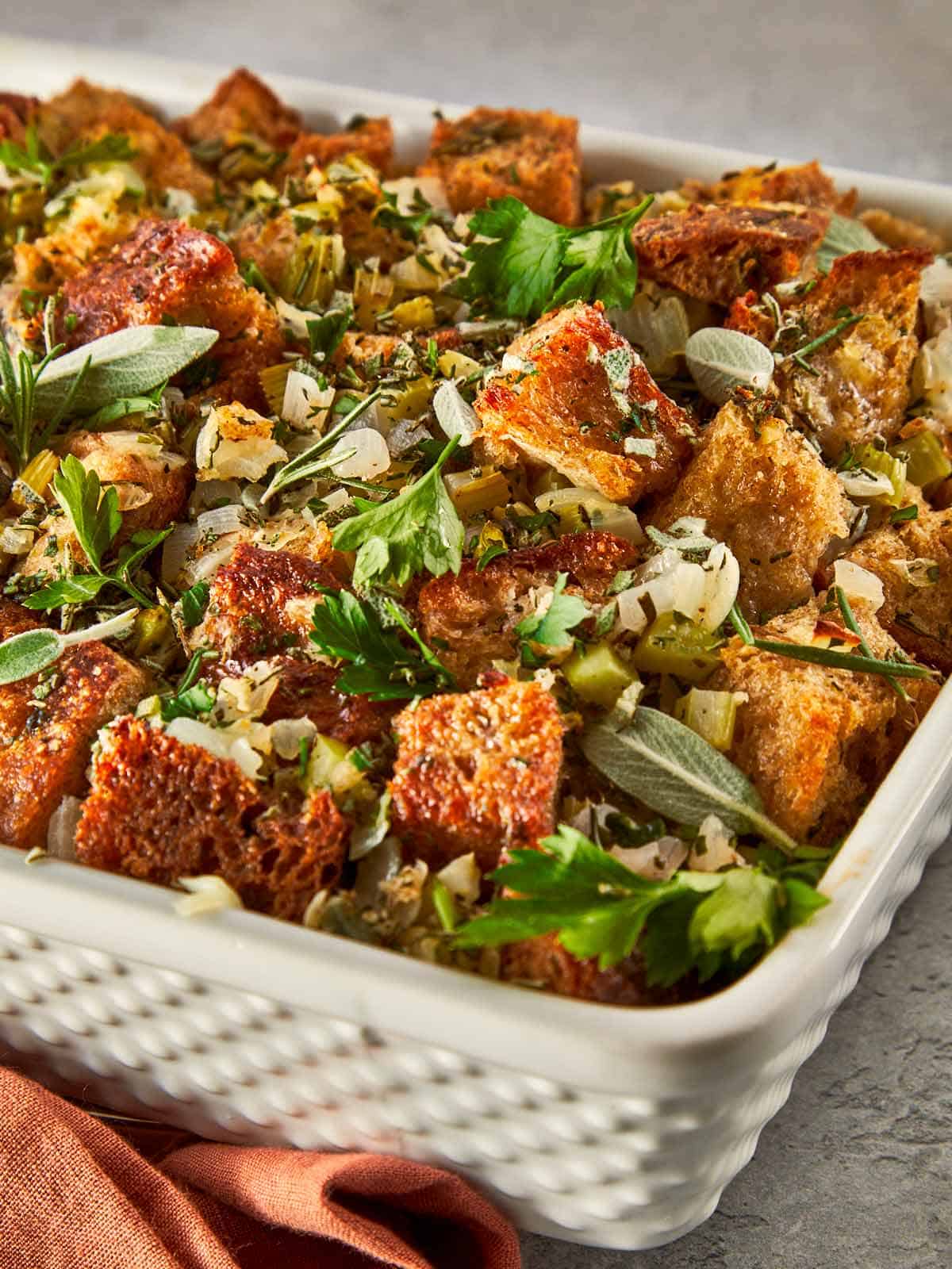 Close-up of a baked herb stuffing with golden bread cubes, sautéed celery and onion, and fresh herbs scattered on top, ready to serve straight from the dish.