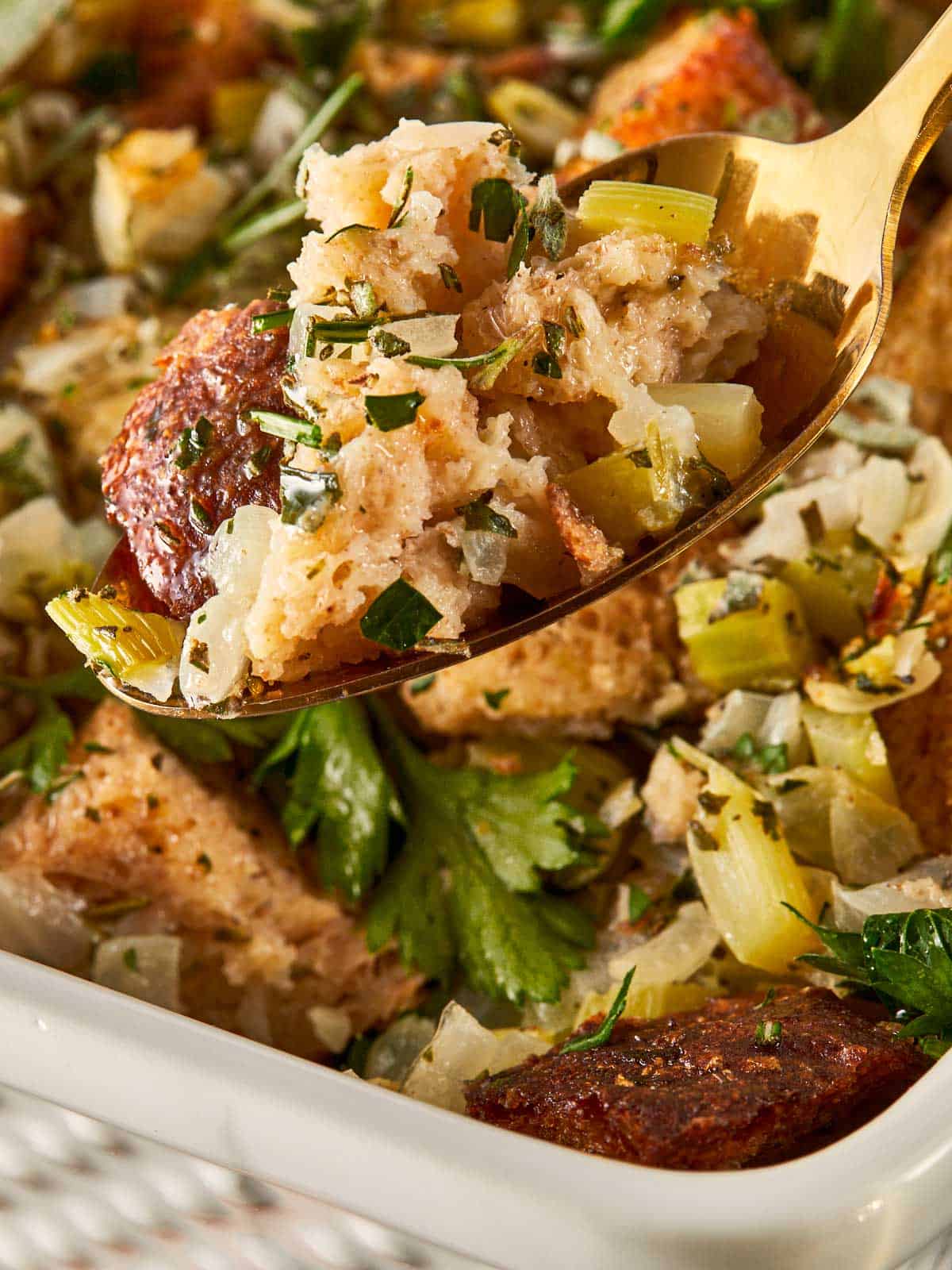 Golden cubes of buttery herb stuffing in a white baking dish, topped with fresh parsley, sage, and rosemary. A gold spoon lifts a perfect bite showing the soft center and crisp edges.