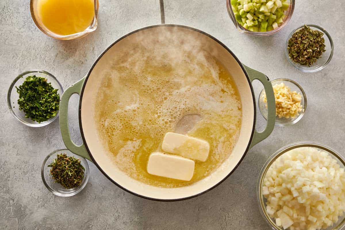 Melting butter in a large green pot, surrounded by bowls of chopped onions, celery, herbs, and broth for classic stuffing.