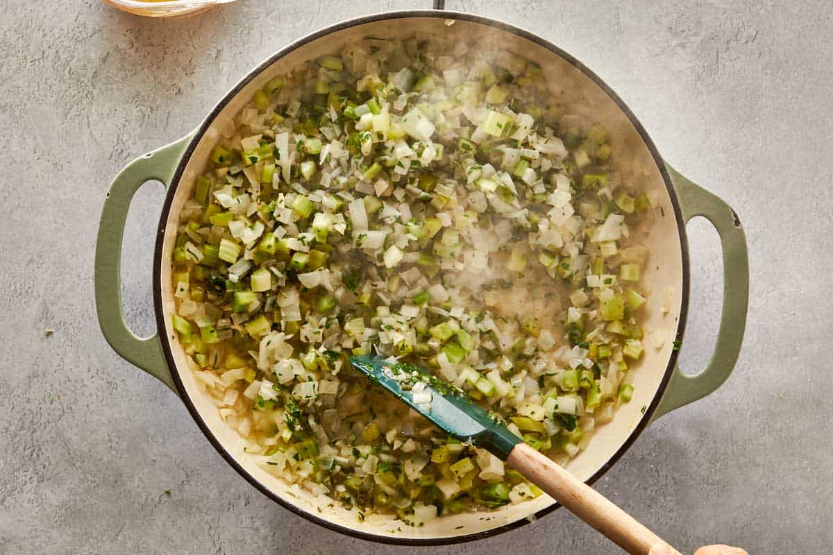 Sautéing chopped onions and celery in a pot with a spatula, with bowls of fresh and dried herbs nearby for stuffing.