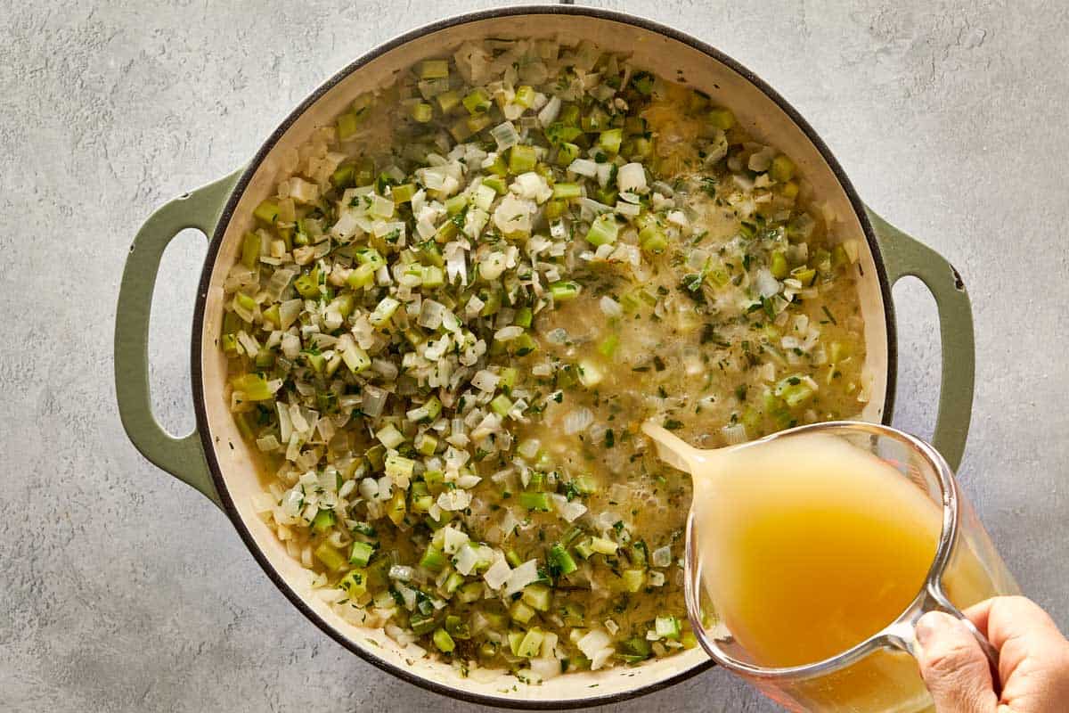 Pouring chicken stock into the pot with sautéed onions, celery, and herbs for moistening stuffing mix.