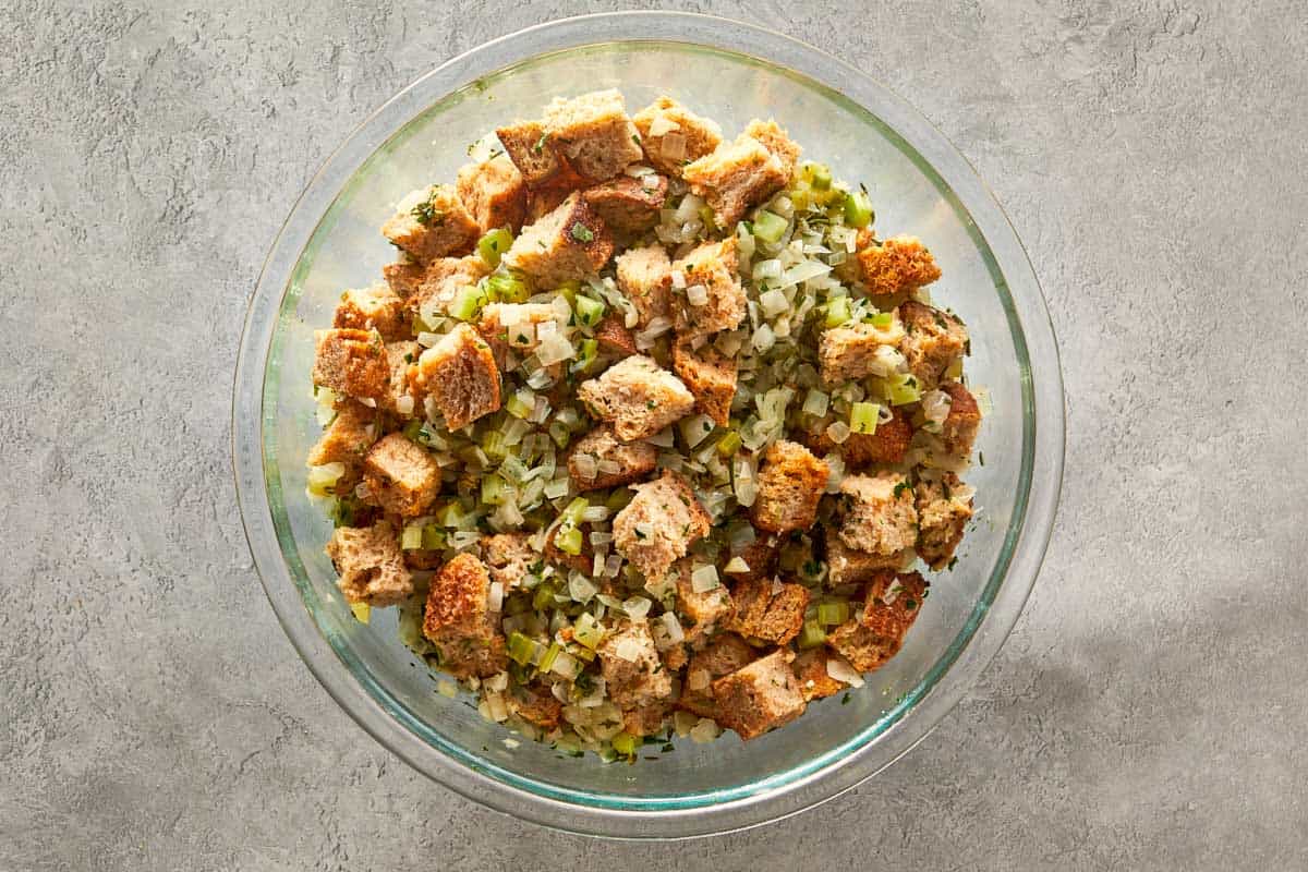 Mixing toasted bread cubes with the cooked onion, celery, and herb mixture in a large glass bowl for homemade stuffing.