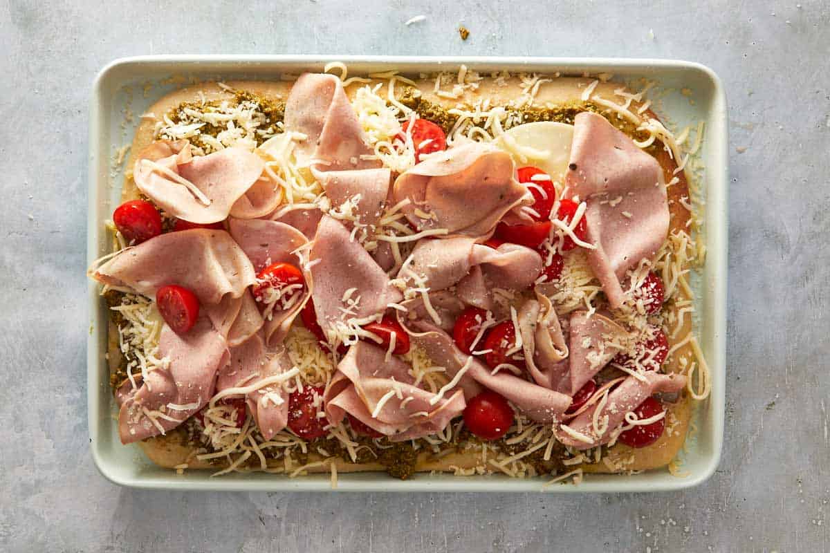 Unbaked pesto focaccia pizza with mortadella, cherry tomatoes, and cheese before going into the oven