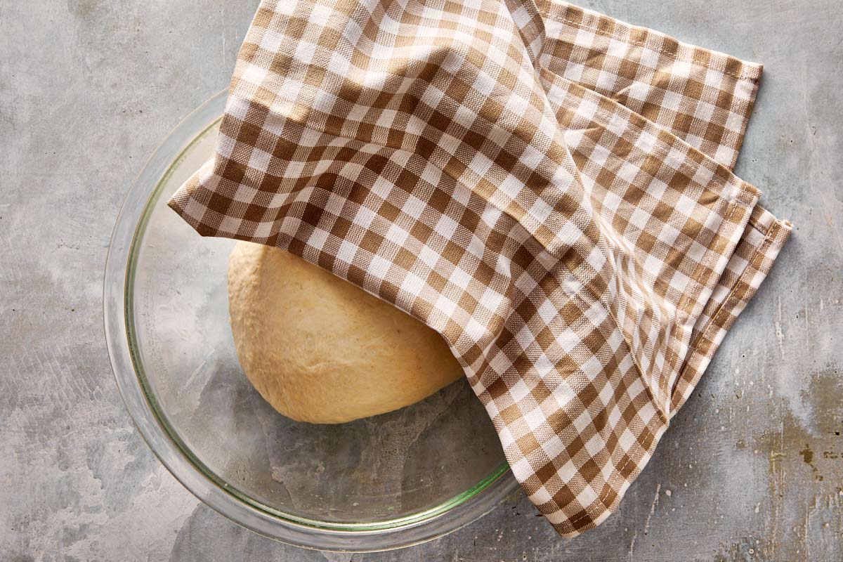 Focaccia dough resting in a bowl, covered with a brown checkered cloth during first rise.