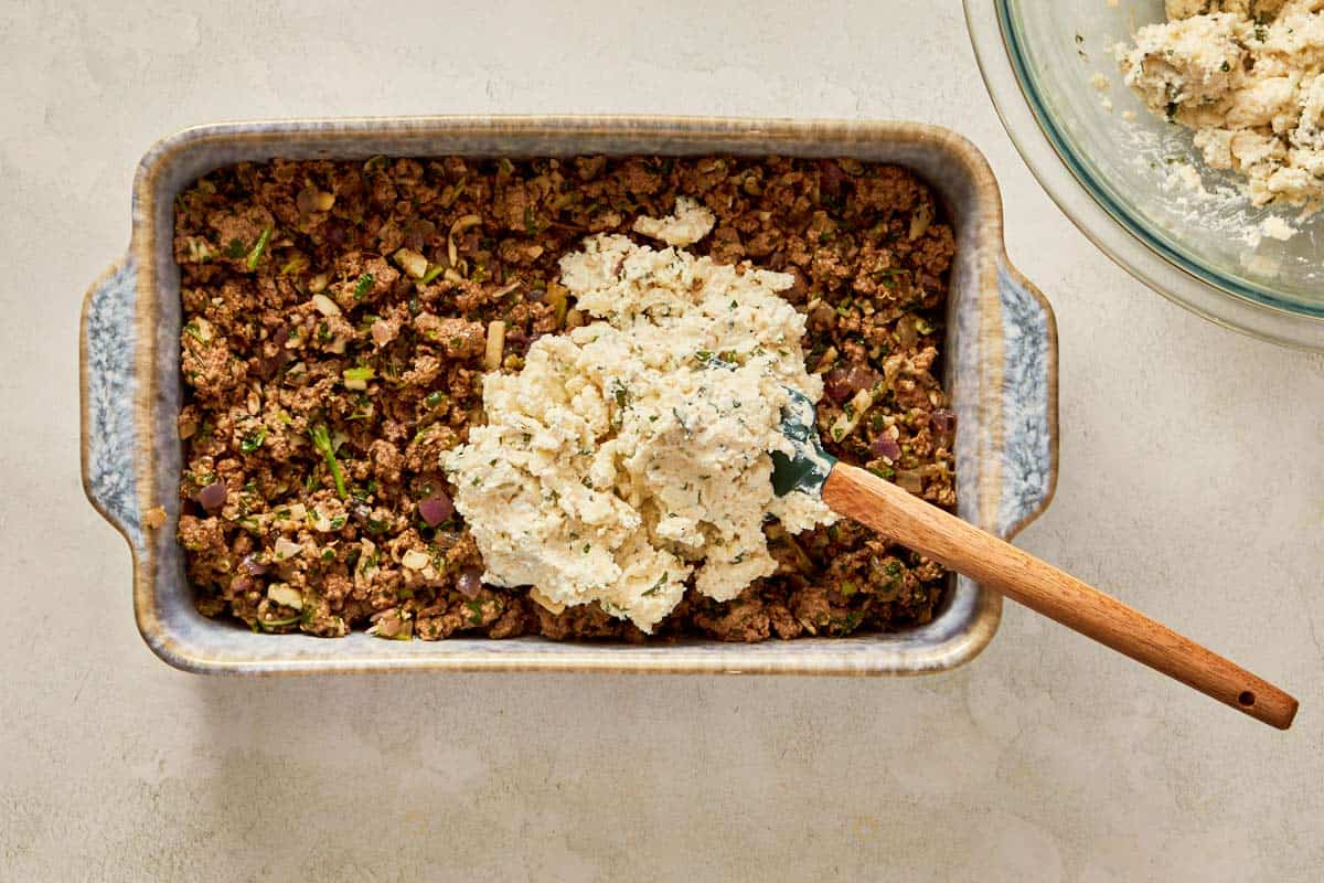 Spreading cheese mixture over cooked beef in a baking dish.