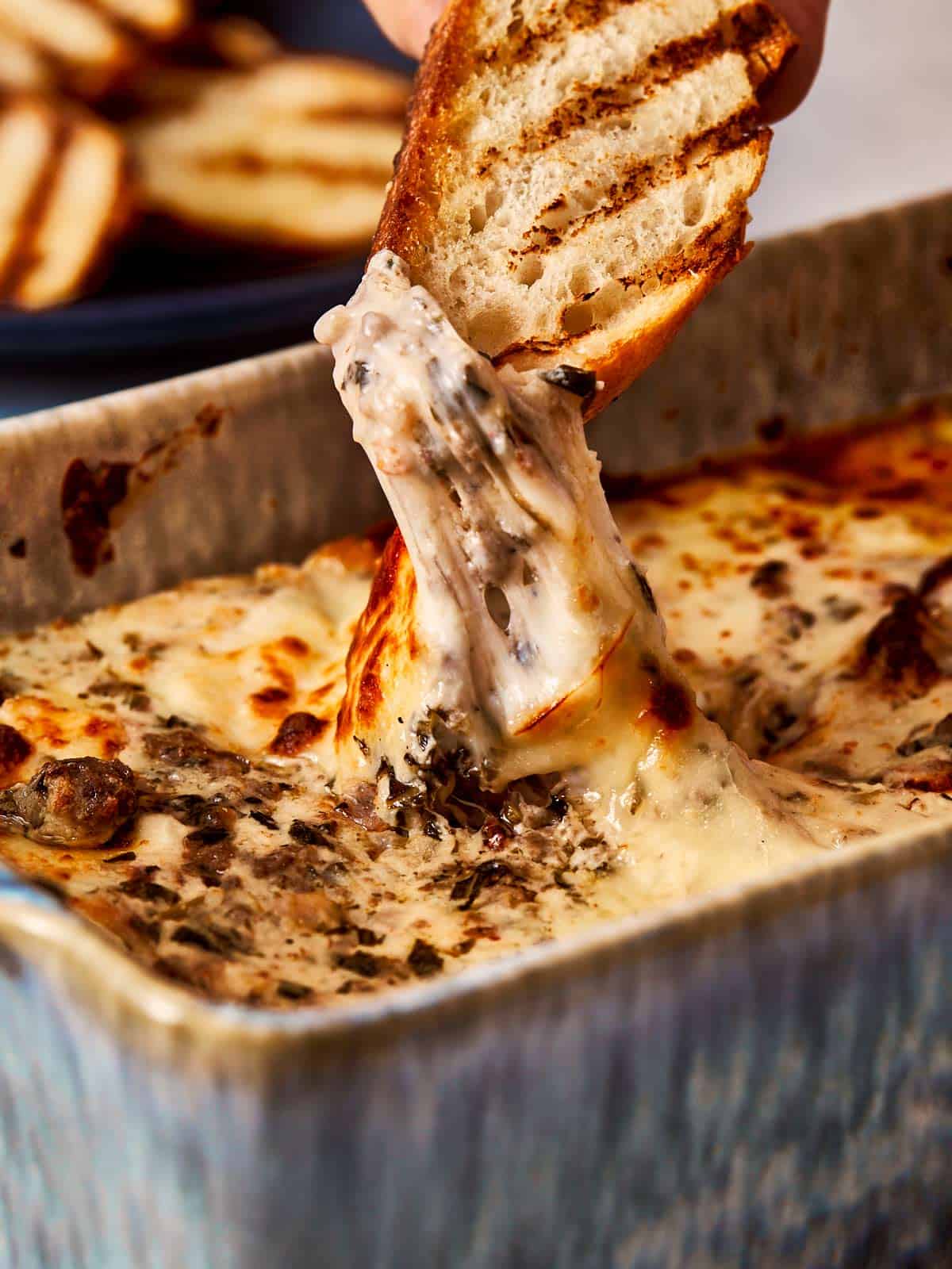 A slice of toasted bread being dipped into a hot, cheesy Lasagna 4 Cheese Dip, showing a gooey stretch of melted cheese with bits of herbs and beef in a golden, bubbling sauce.