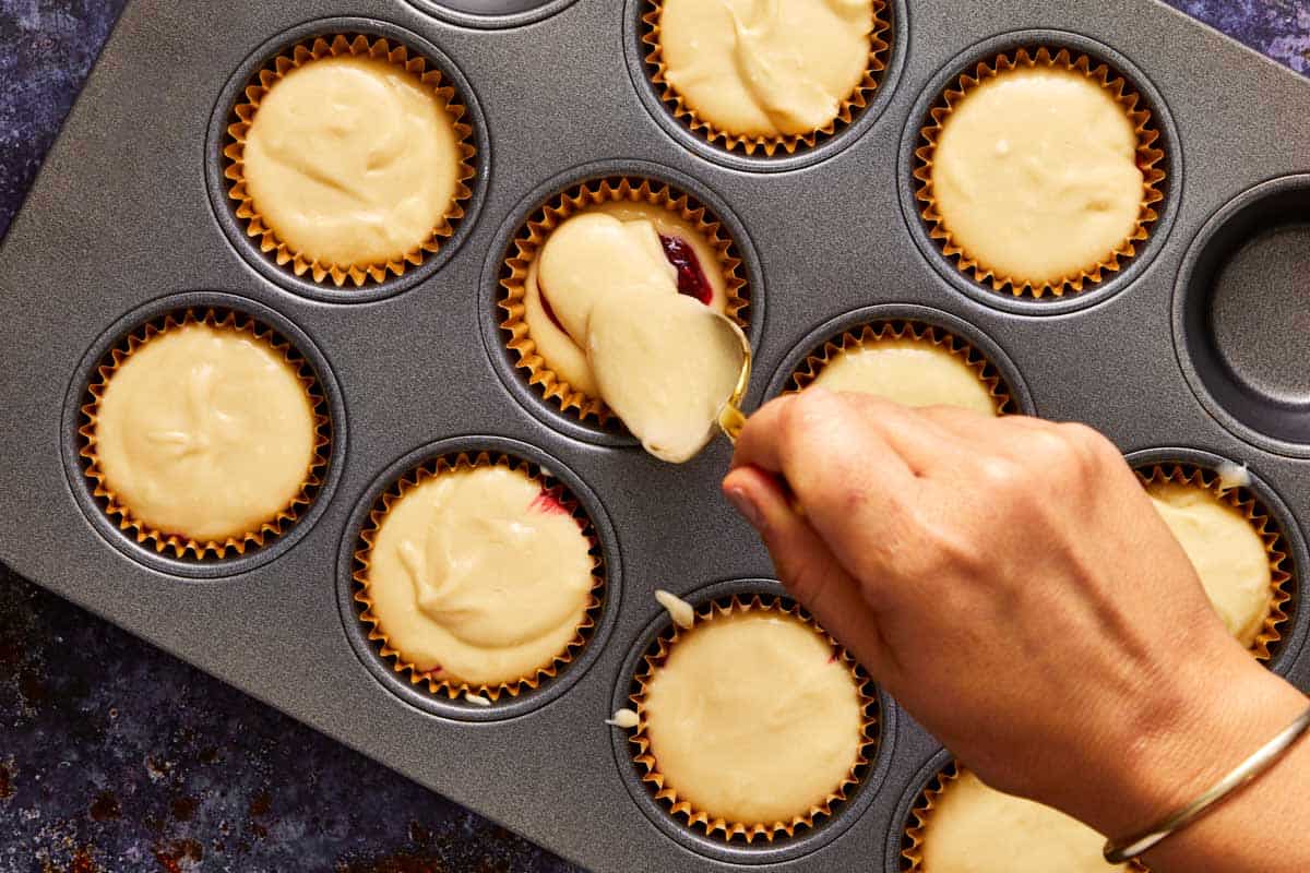 Hand spooning muffin batter into paper liners, adding a frozen raspberry insert in the center of each muffin tin.