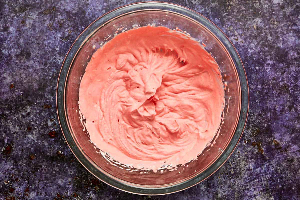 Bowl of pink whipped cream topping, made with heavy cream, vanilla, and red food coloring, ready for piping.