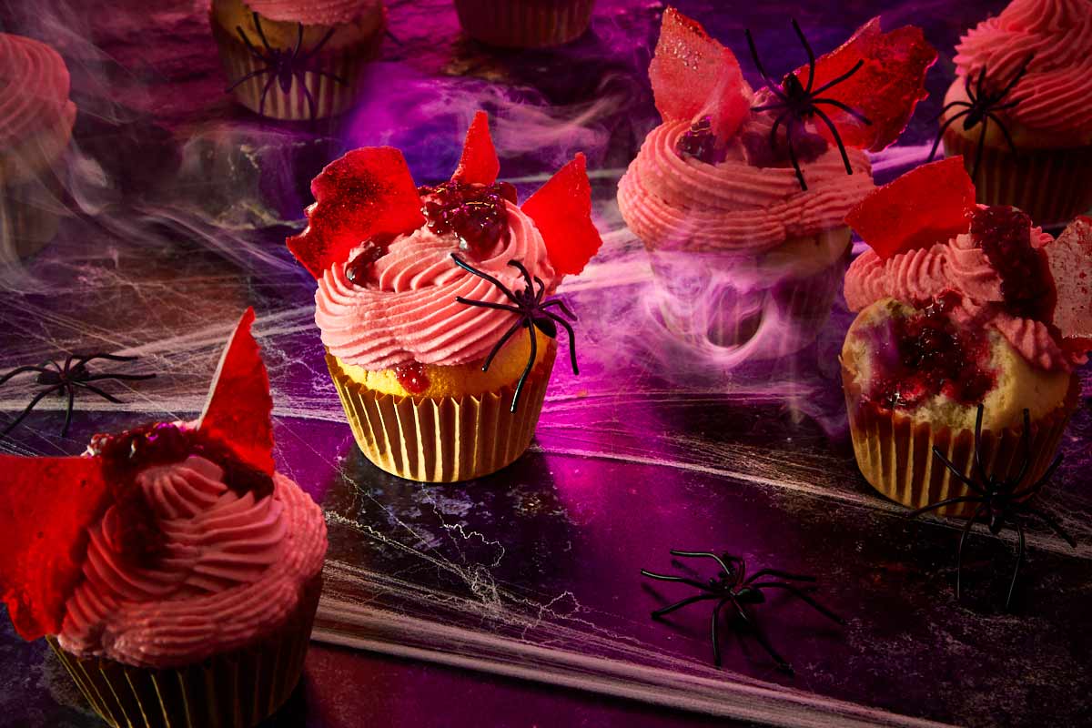 Halloween scene with frosted raspberry muffins decorated with fake spiders, candy glass shards, and foggy purple lighting.