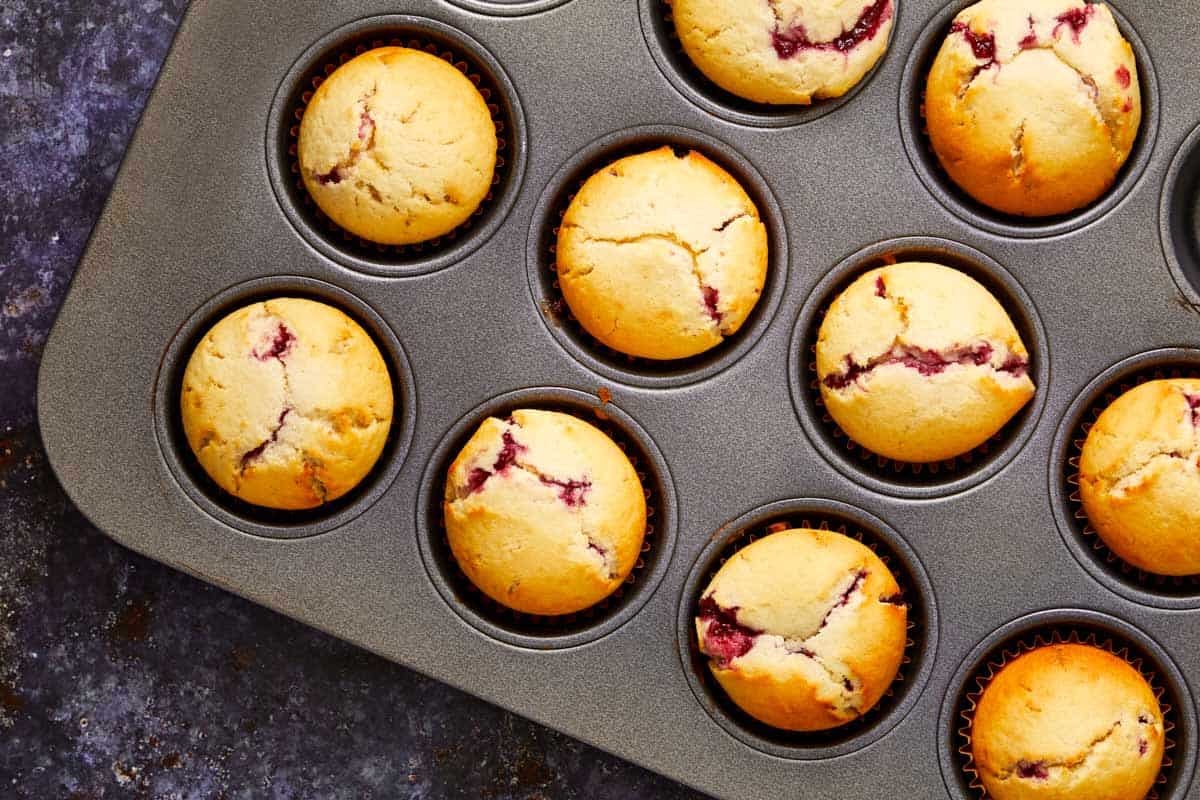 Freshly baked raspberry muffins cooling in a muffin tin, golden on top with red raspberry filling peeking through the cracks.