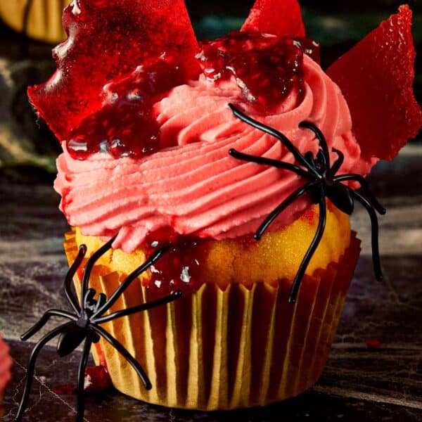 Close-up of a Halloween raspberry muffin topped with pink whipped cream, raspberry coulis, and broken glass candy shards, decorated with fake black spiders.