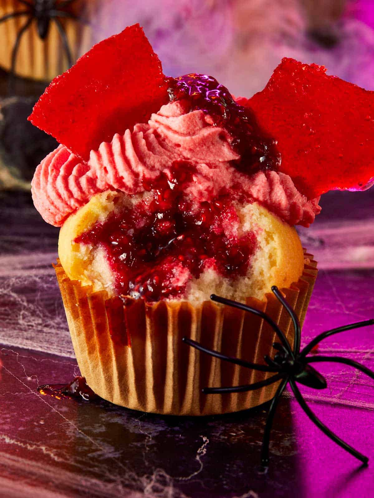 Raspberry muffin cut open to reveal a red raspberry filling, topped with whipped cream, candy glass shards, and a fake black spider on a spooky purple surface.