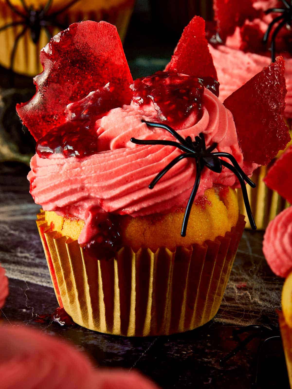 Close-up of a Halloween raspberry muffin topped with pink whipped cream, raspberry coulis, and broken glass candy shards, decorated with fake black spiders.