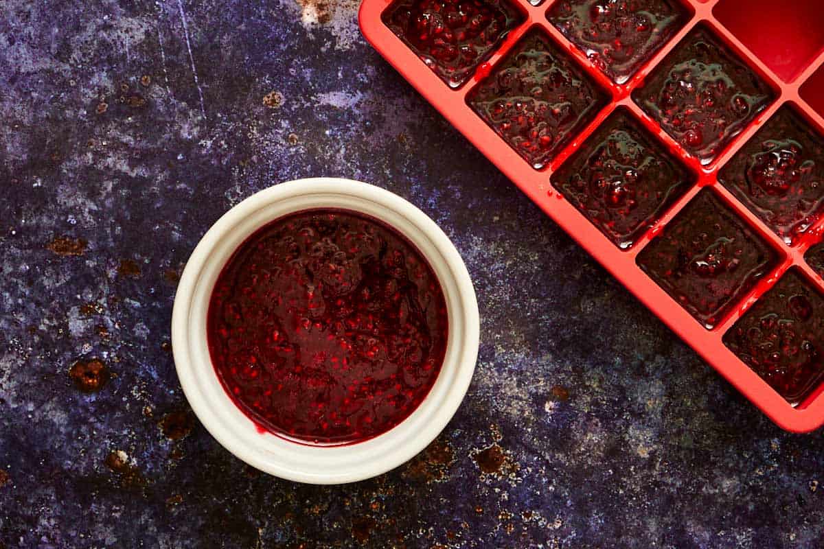 Homemade raspberry coulis divided into a small bowl and an ice cube tray, ready to freeze for the muffin filling.