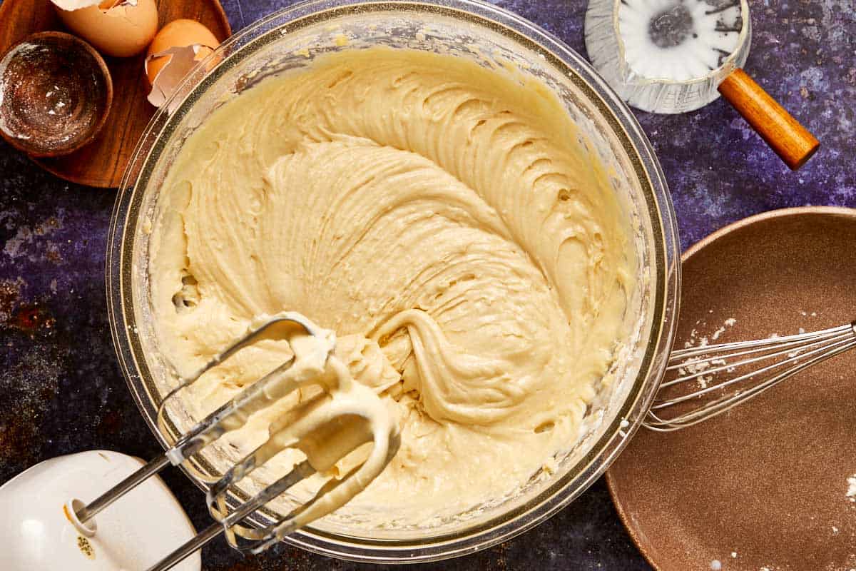 Mixing bowl filled with smooth muffin batter being whipped with an electric mixer, surrounded by eggs and baking tools.