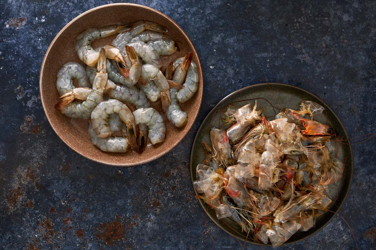 Peeled raw shrimp in one bowl and shrimp shells and heads in another, ready to make broth.