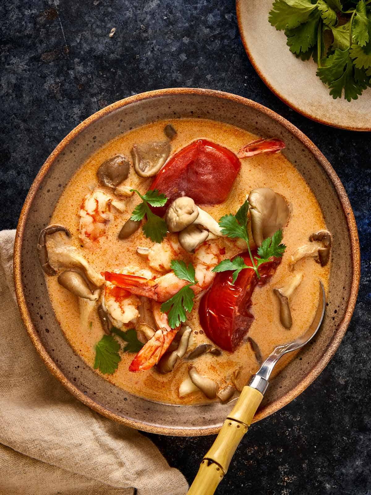 A warm bowl of Tom Yum Kha Soup with shrimp, mushrooms, and tomatoes in a creamy coconut broth, garnished with fresh cilantro.