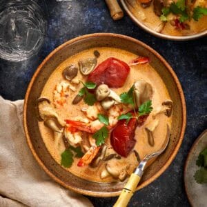 Overhead shot of creamy Thai Tom Yum Kha soup with shrimp, mushrooms, and tomatoes served in a rustic bowl with a bamboo-handled spoon and fresh cilantro on top.
