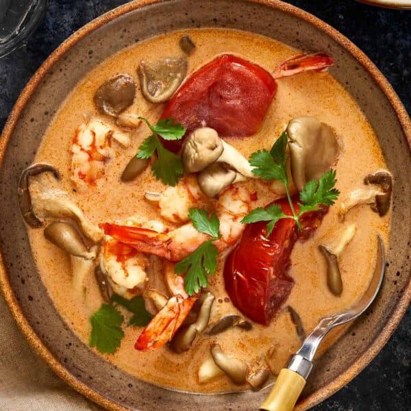 Overhead shot of creamy Thai Tom Yum Kha soup with shrimp, mushrooms, and tomatoes served in a rustic bowl with a bamboo-handled spoon and fresh cilantro on top.