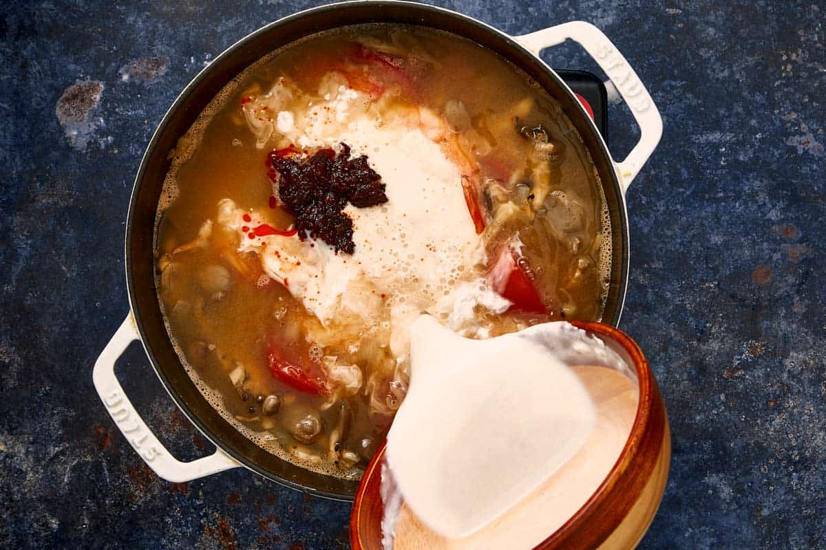 Pouring coconut milk and Thai chili paste into a pot of simmering soup.