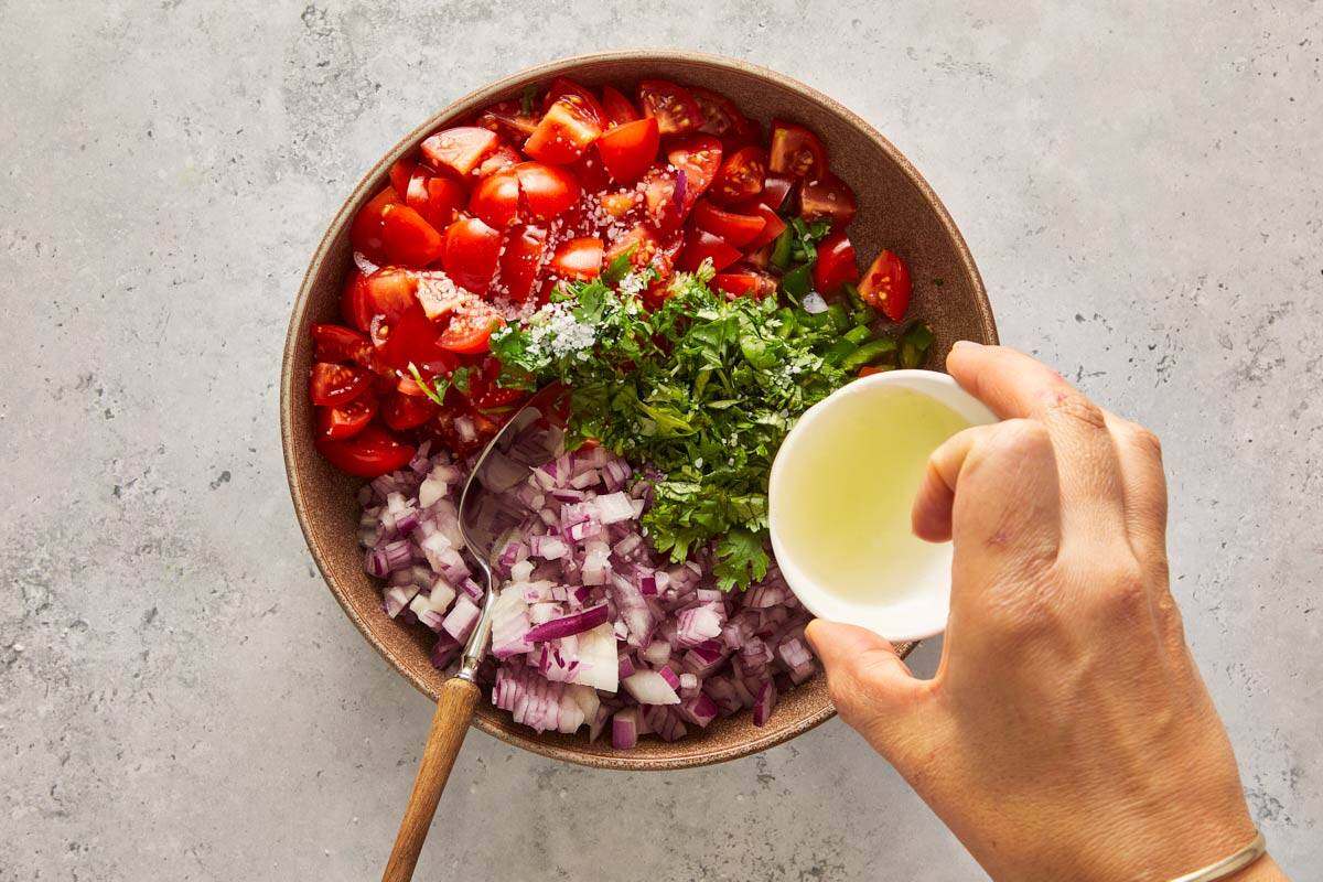 Hand pouring lime juice over chopped tomatoes, onions, and cilantro in a bowl