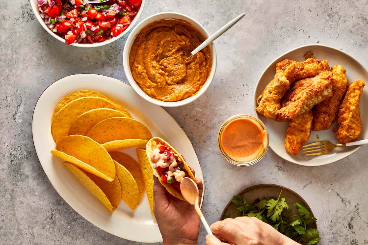 Hands assembling crispy fish tacos with pico de gallo, sauce, and taco shells on the table
