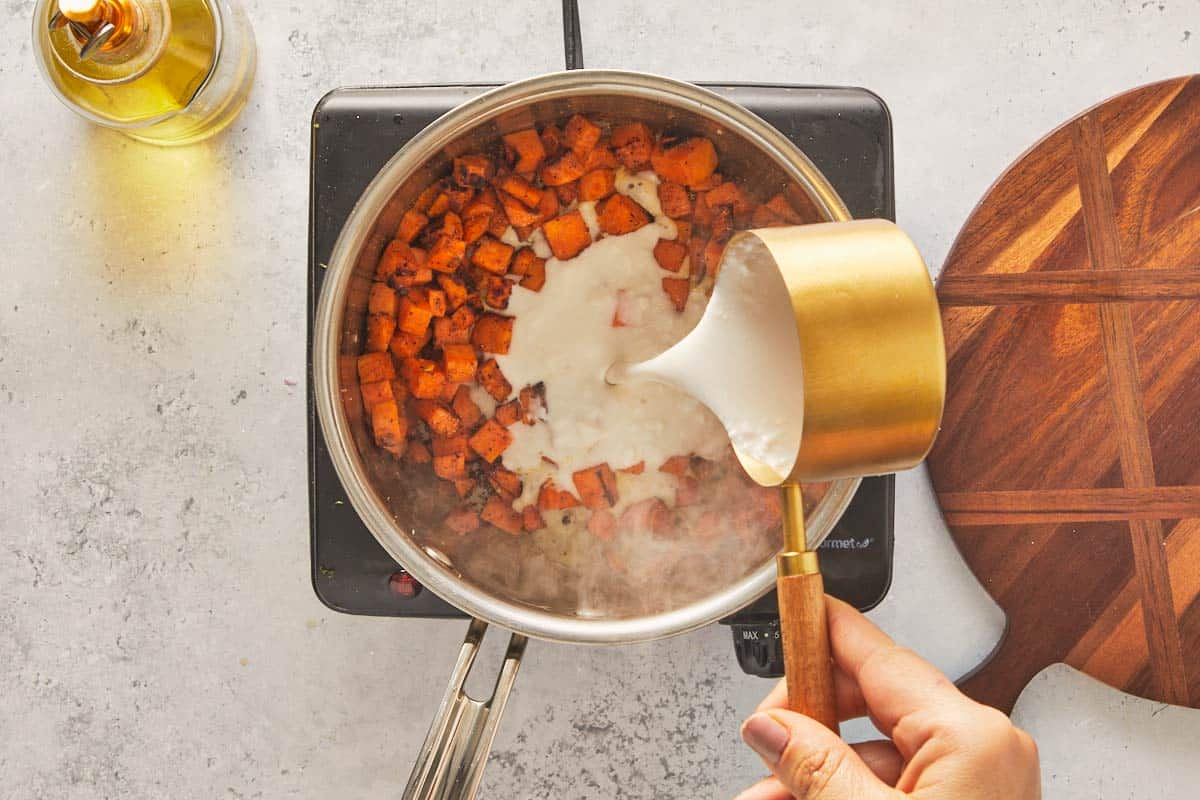 Hand pouring coconut cream into a saucepan with cooked sweet potato cubes
